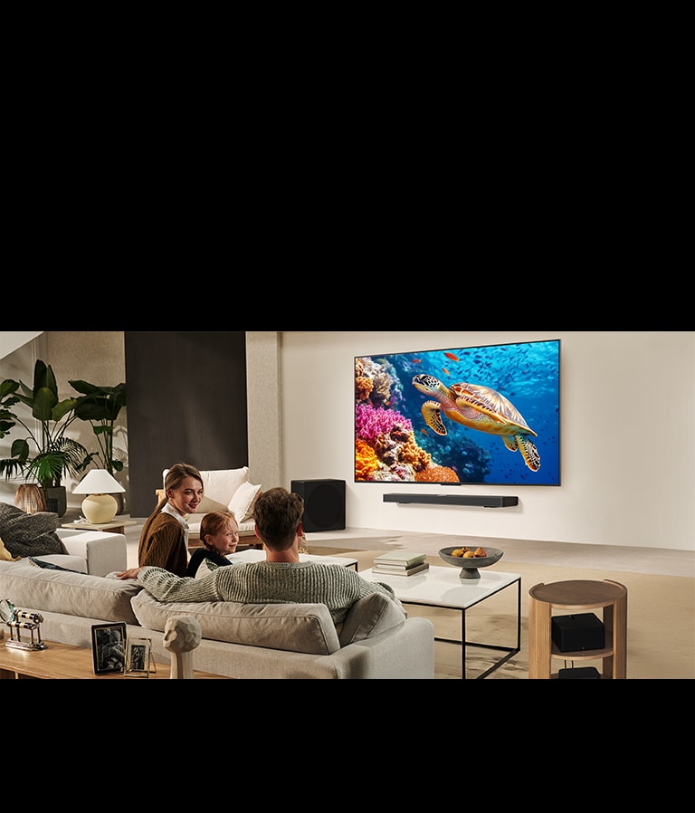 Šeima sėdi ant sofos priešais LG TV, sumontuotą ant sienos virš „LG Soundbar“. „Zero Connect Box“ yra kavos staliuko viduje.