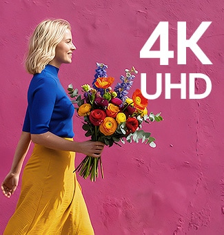 Mobilusis ekranas „LG StanbyME 2 Max LX6“ su 4K UHD raiška rodo spalvingais drabužiais vilkinčią moterį, laikančią gėles, einančią pro violetinę gatvės sieną, išryškindamas ryškią ir detalią raišką.