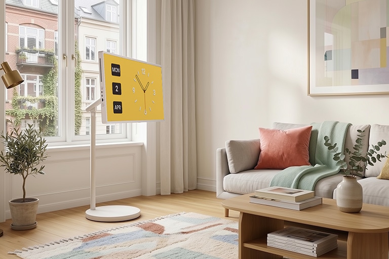 „LG StanbyME 2 Max LX6“ kilnojamasis ekranas su „Roll & Carry“ dizainu stovi prie lango kompaktiškame, balto atspalvio kambaryje su laikrodžio ekranu, pabrėžiančiu lankstų išdėstymą ir lengvą mobilumą universaliam naudojimui.