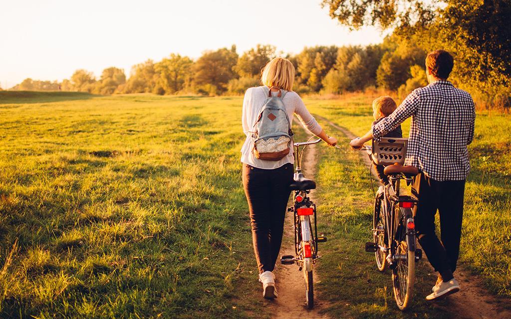 Two parents and a child in a family walking their bicycles in a paddock near the forest