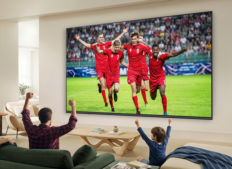 LG QNED evo AI QNED80 Mini LED Ultra Big TV uzstādīts pie sienas gaišā viesistabā, kurā redzama futbola svinēšanas aina ar dzīvīgām krāsām un izsmalcinātu attēla kvalitāti plašā ekrānā, kamēr uz dīvāniem sēdoša ģimene kopā priecājas.