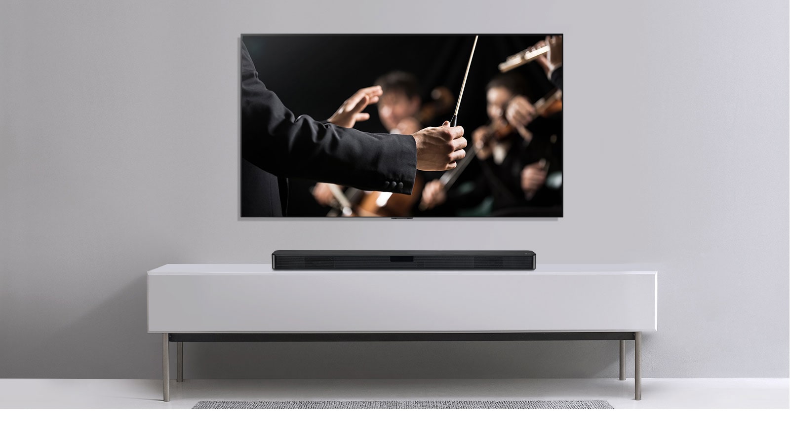 Pie pelēkas sienas redzams televizors, un zem tā uz pelēka plaukta novietota LG skaņas josla. Televizorā redzams diriģents ar orķestri.