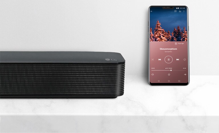 Tuvplāns ar LG Soundbar labajā pusē blakus viedtālrunim. Baltajā plauktā atrodas divas ierīces.