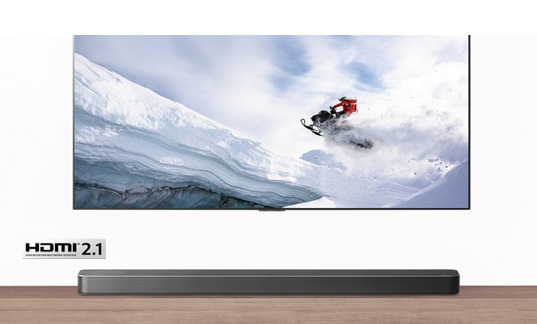 TV un skaņas josla no priekšas. TV redzams vīrietis uz sniega motocikla sniegotos kalnos. HDMI 2.1 logotips zem TV.