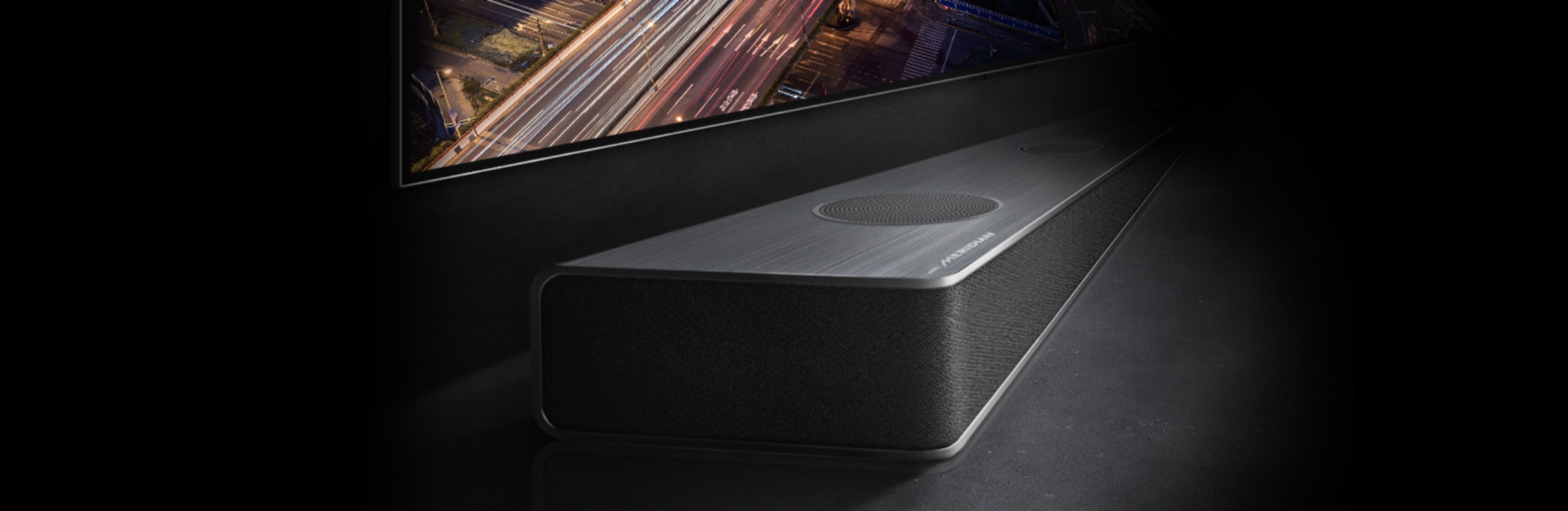 Zem televizora ekrāna, kurā attēlots nakts skats uz ceļu, redzama neuzkrītoša soundbar mala.