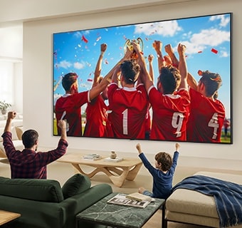 LG QNED evo AI QNED85 Mini LED Ultra Big TV uzstādīts uz sienas viesistabā, kurā redzama sporta uzvaras svinēšana ar dzīvīgām krāsām un izsmalcinātu attēla kvalitāti plašajā ekrānā, ģimenei kopā sēžot dīvānos un priecājoties.