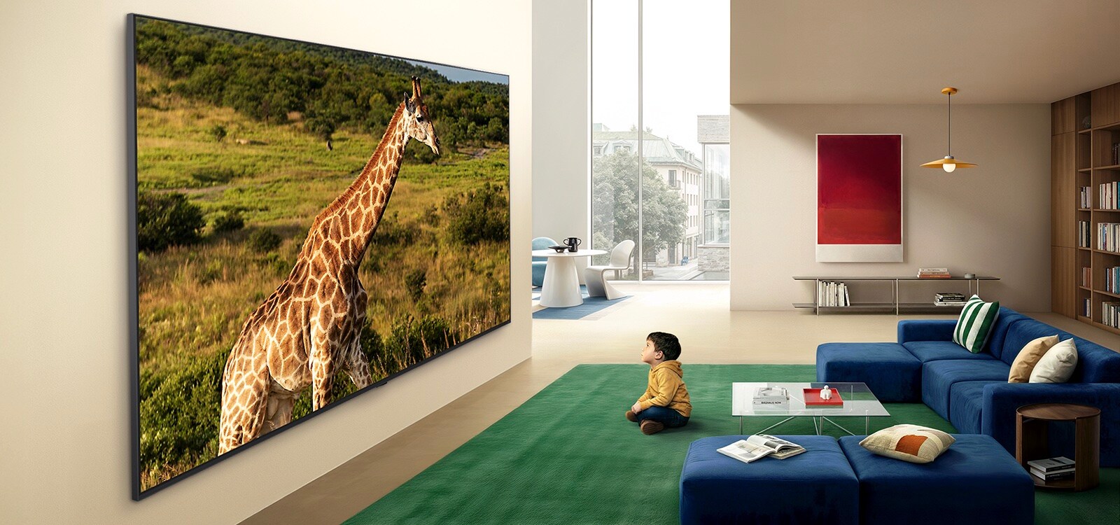 LG QNED evo AI QNED85 Mini LED Ultra Big TV uzstādīts pie sienas gaišā dzīvojamā telpā, kurā redzama žirafe dabas ainavā ar dzīvīgām detaļām, kas uzsver aizraujošo mērogu un efektu bērnam, kurš sēž tuvumā.