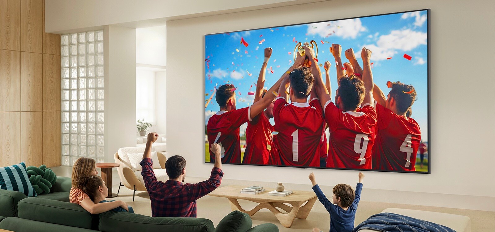 LG QNED evo AI QNED85 Mini LED Ultra Big TV uzstādīts uz sienas viesistabā, kurā redzama sporta uzvaras svinēšana ar dzīvīgām krāsām un izsmalcinātu attēla kvalitāti plašajā ekrānā, ģimenei kopā sēžot dīvānos un priecājoties.