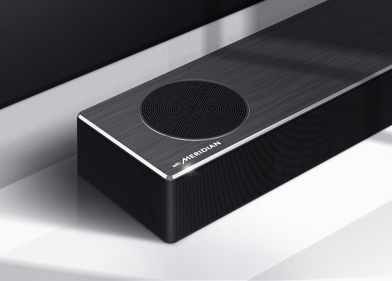 Tuvplānā redzama LG skaņas joslas kreisā puse ar Meridian logotipu produkta apakšējā kreisajā stūrī. Redzama arī televizora apakšējā kreisā puse.