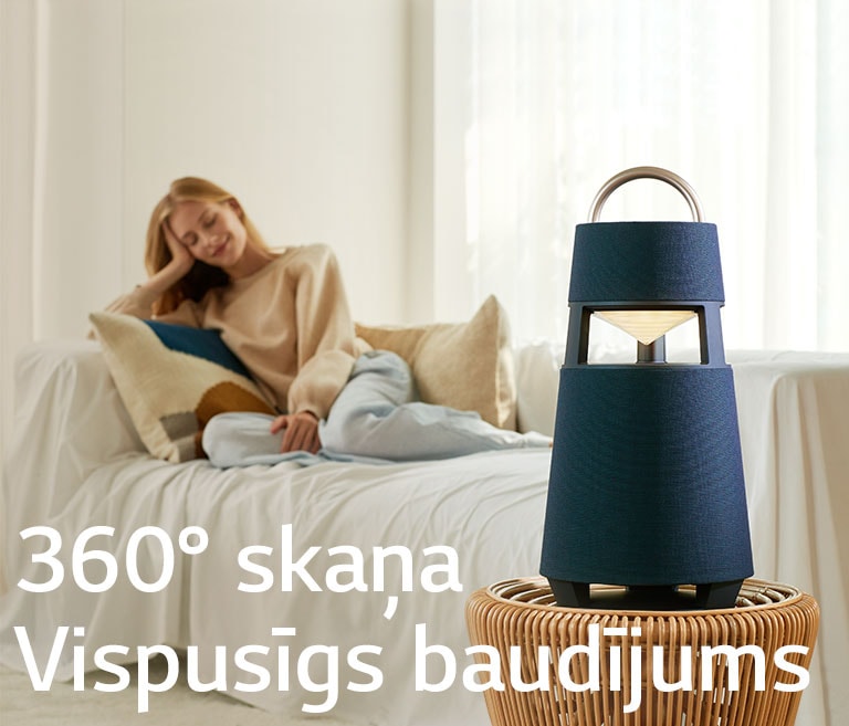 Attēlā redzama sieviete, kas sēž dīvānā un klausās mūziku no pretī stāvošā XBOOM 360.