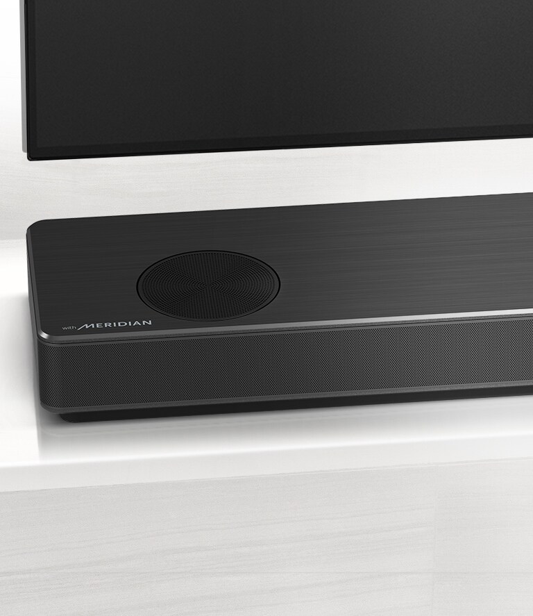 Pietuvinājumā redzama LG skaņas joslas kreisā puse ar Meridian logotipu apakšējā kreisajā stūrī. Redzama arī televizora apakšējā kreisā puse.