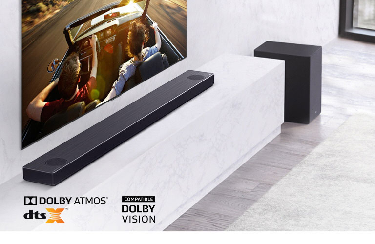 Televizors pie sienas, LG skaņas josla zem tā uz balta marmora plaukta un zemo frekvenču pastiprinātājs labajā pusē. Televizorā redzams pārītis braucam mašīnā.