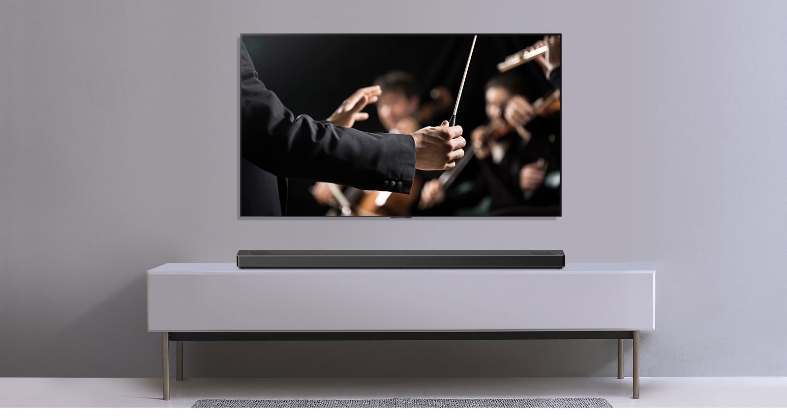 Pie pelēkas sienas redzams televizors, un zem tā uz pelēka plaukta novietota LG skaņas josla. Televizorā redzams diriģents ar orķestri.