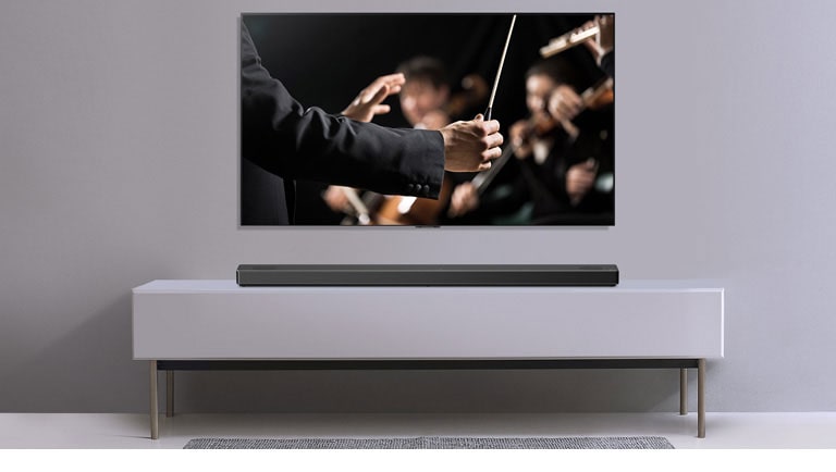 Pie pelēkas sienas redzams televizors, un zem tā uz pelēka plaukta novietota LG skaņas josla. Televizorā redzams diriģents ar orķestri.