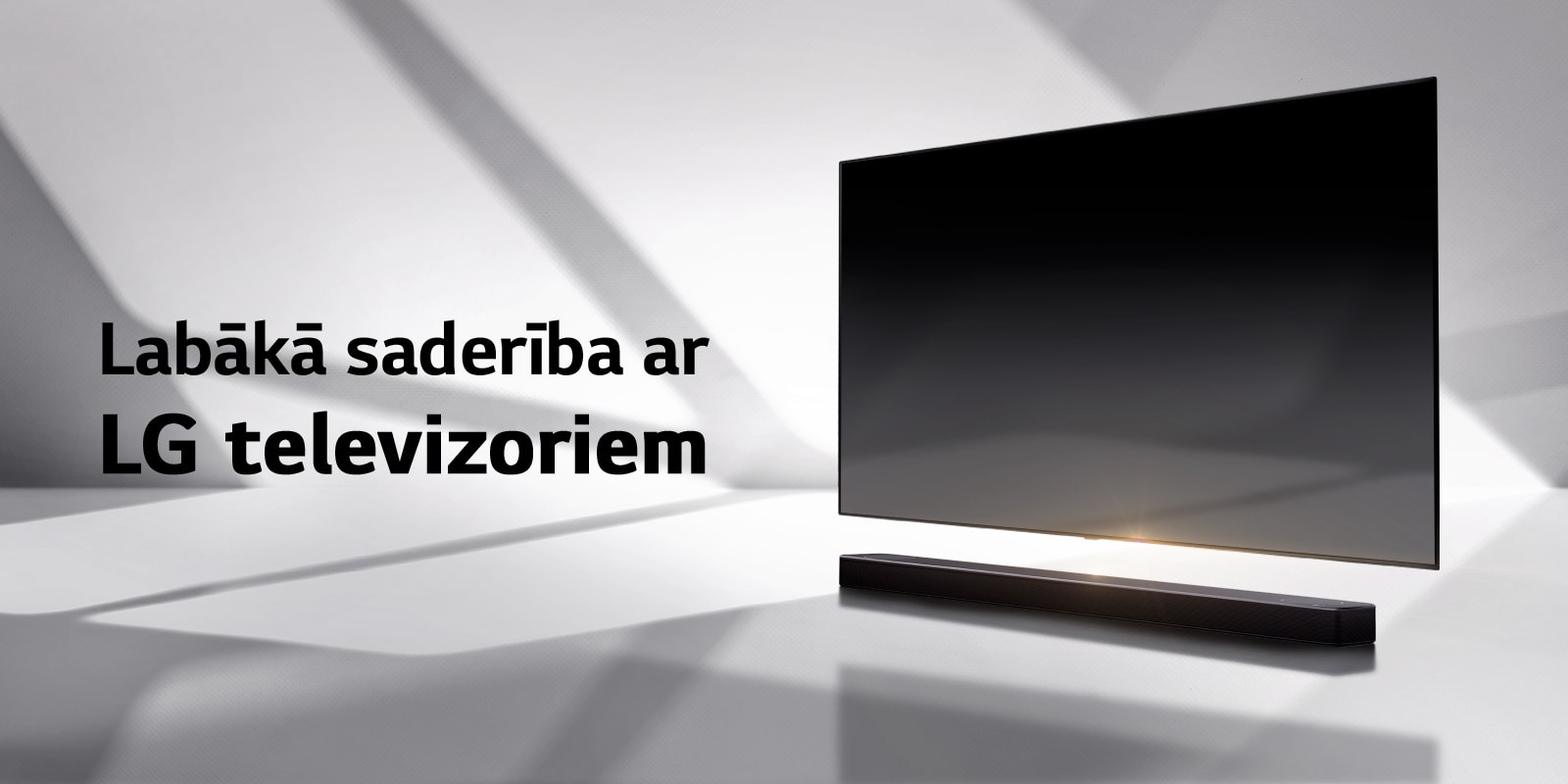 Skaņas josla un televizors novietoti uz baltas grīdas, un no ārpuses aiz tiem stiepjas ēna.
