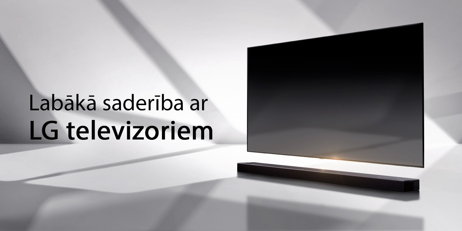 Skaņas josla un televizors novietoti uz baltas grīdas, un no ārpuses aiz tiem stiepjas ēna.