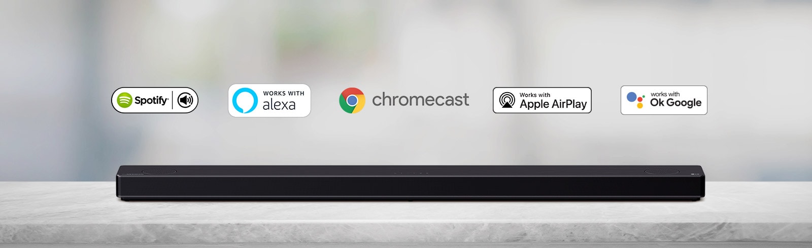 Uz pelēka plaukta stāv skaņas josla un no labās uz kreiso pusi ir redzami sarindoti AI platformu logotipi — Spotify, Alexa, Chromecast, Apple Airplay un OK Google.