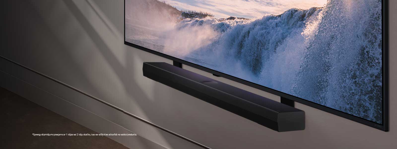 LG TV un Soundbar ir novietoti leņķiskā perspektīvā un piestiprināti pie sienas. TV tiek parādīts liela ūdenskrituma tuvplāns un maiga saules gaisma kaskāde pāri sienai, TV un Soundbar. Atruna: “Synergy stiprinājums pieejams ar 1 kājas vai 2 kāju statīvu, kas var atšķirties atkarībā no valsts/produkta.”