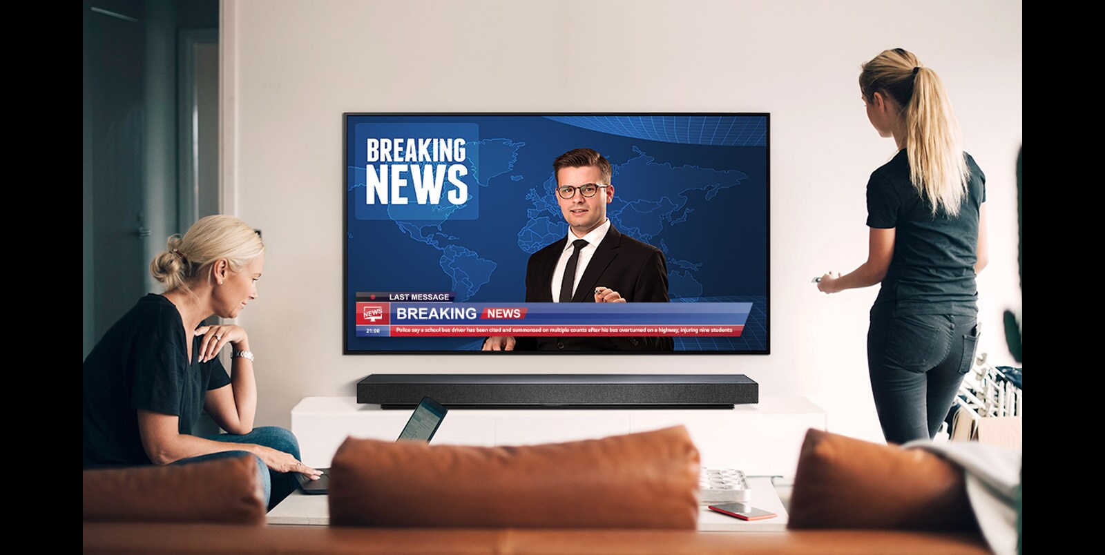 Sieviete televizorā skatās ziņas maksimālā kvalitātē, stāvot kājās, un otra sieviete sēž pie sava klēpjdatora (pārvietojiet slīdni)