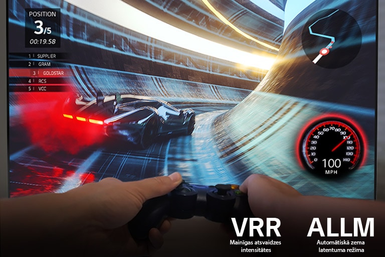 Ātrumsacīkšu spēles ekrāns un iepretim divas rokas, kas tur spēļu vadības pulti 