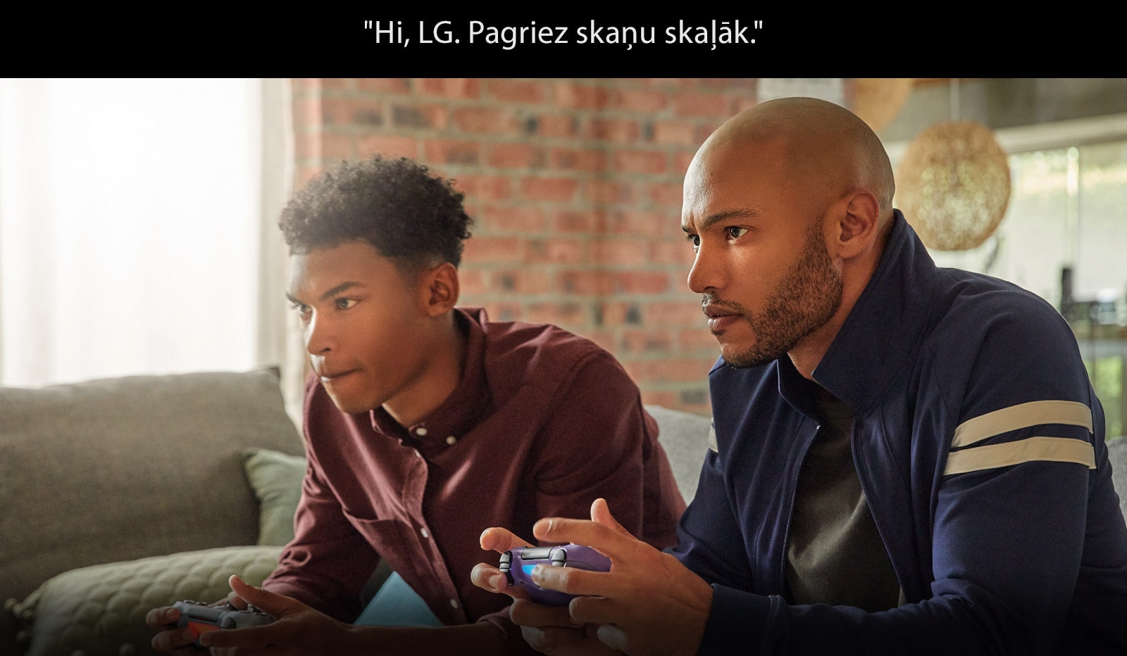 Divi vīrieši ar bezvadu spēles vadības pultīm sēž dīvānā un ir iegrimuši spēlē