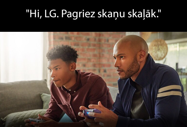 Divi vīrieši ar bezvadu spēles vadības pultīm sēž dīvānā un ir iegrimuši spēlē