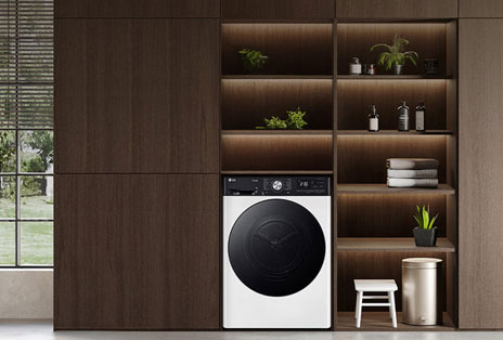 LG Tumble Dryer baltā krāsā ar melnām durvīm, ievietots tumša koka plauktos ar zaļiem augiem un salocītiem dvieļiem.