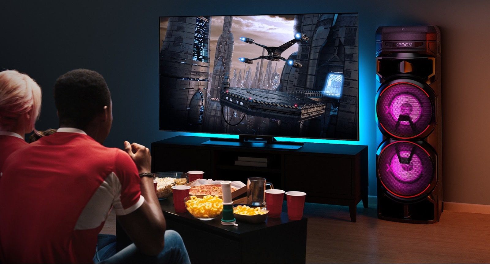 Cilvēki skatās pie sienas piestiprinātu televizoru ar LG XBOOM RNC7 pa labi no tā. Televizora ekrānā tiek rādīta filma.