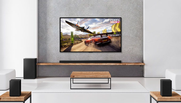 Baltā viesistabā redzami divi aizmugures skaļruņi, zemo frekvenču pastiprinātājs, Soundbar un televizors. Televizora ekrānā redzama braucoša automašīna un kosmosa kuģis.