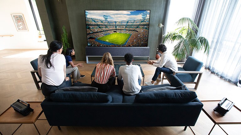 Cilvēki skatās sporta pārraidi viesistabas televizorā ar pieslēgtiem Bluetooth aizmugures skaļruņiem