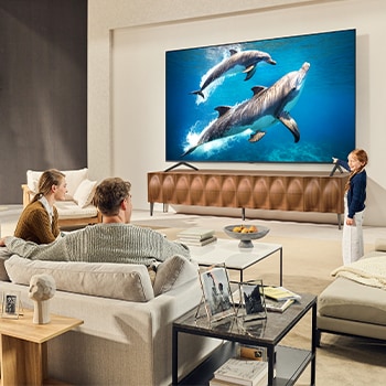 LG 65 collu LG QNED evo AI MiniLED 4K viedtelevizors QNED93 2025, Ģimene sēž uz dīvāna pretī LG QNED TV, kas sēž virs TV statīva, un maza meitene norāda uz ekrānu, kurā attēloti divi delfīni. Virsrakstā norāde par to, kā iespējams baudīt nevainojamu izšķirtspēju lie, 65QNED93A6A, thumbnail 13