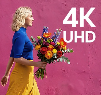 LG StanbyME 2 Max LX6 pārvietojamajā ekrānā ar 4K UHD izšķirtspēju redzama sieviete krāsainā apģērbā, kura tur ziedus, pastaigājoties garām violetai ielas sienai, izceļot asu un detalizētu izšķirtspēju.