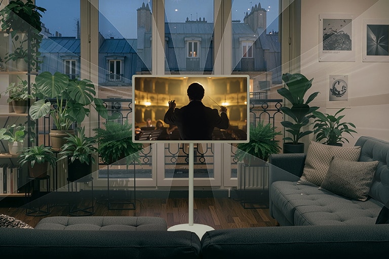 LG StanbyME 2 Max LX6 pārvietojamais ekrāns ar AI Sound Pro tehnoloģiju rāda orķestra ainu augiem piepildītā, tumšā, mājīgā telpā, skaņas viļņiem stiepjoties virzienā uz cilvēku, kurš lasa uz dīvāna, ilustrējot ieskaujošu, telpu piepildošu audio.