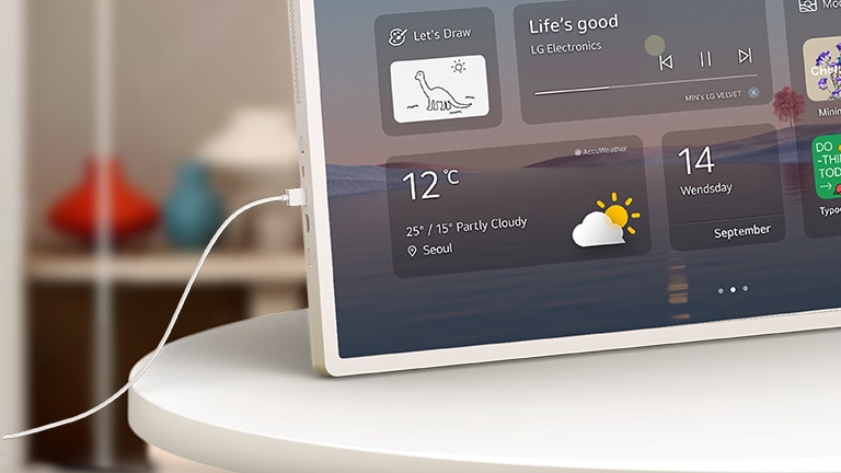 LG StanbyME 2 Max LX6 pārvietojamais ekrāns ar USB-C kabeli ir parādīts uz galda ar kabeli, kas pievienots tā portam, attēlojot mājas logrīka saskarni, izceļot vienlaicīgu uzlādi un ierīču savienojamību.