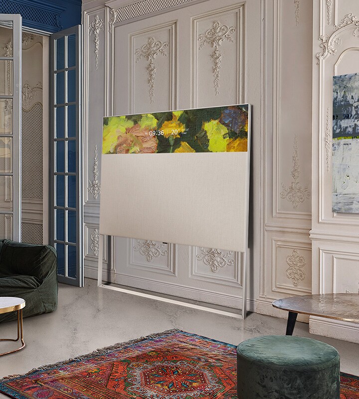 EASEL televizors Line View skatījumā, kas ir atstutēts pret sienu ar dekoratīvu reljefu. Tas atrodas blakus gleznai uz sienas un aiz sarežģīta dizaina paklāja.