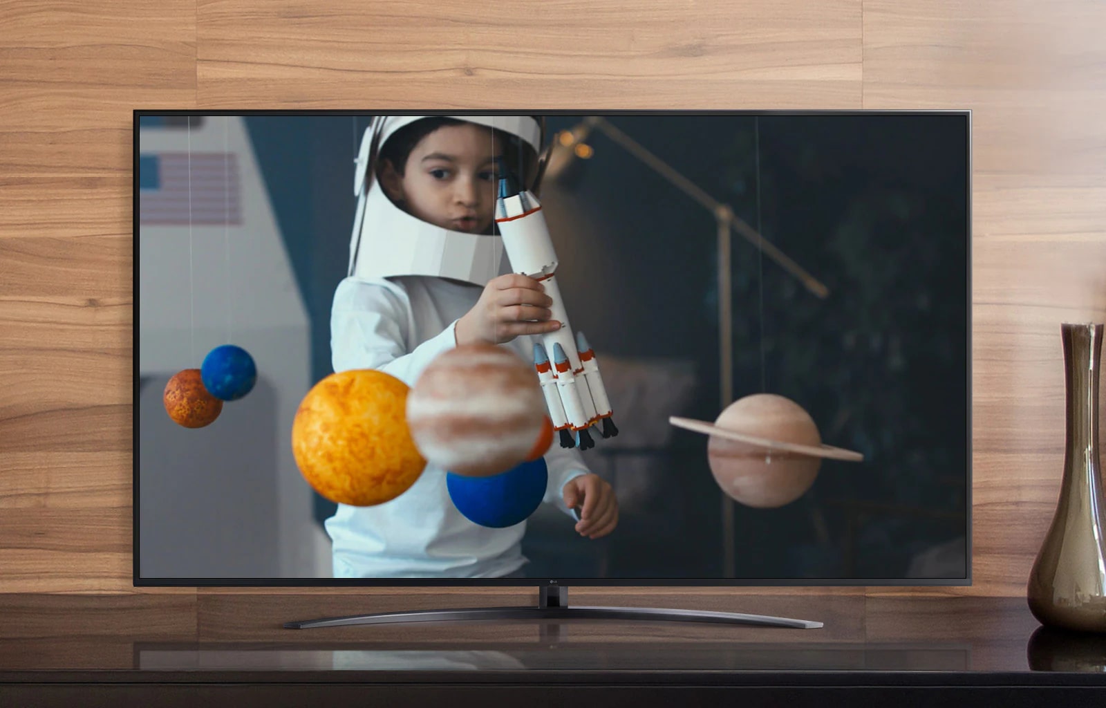 La pantalla de un televisor que reproduce un video de un niño con un traje de astronauta hecho por él, jugando con una nave espacial en su habitación decorada con miniaturas de planetas (reproducir el video)