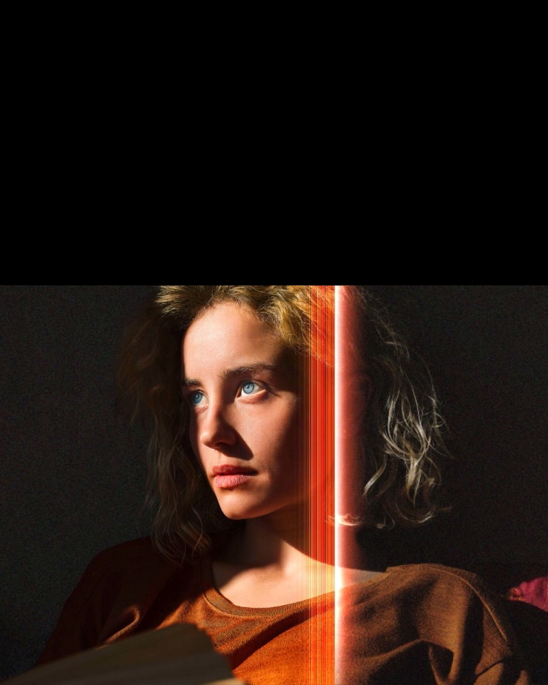 A woman with piercing blue eyes and a burnt orange top in a dark space. Red lines depicting AI refinements cover part of her face, which is bright and detailed, while the rest of the image looks dull. 