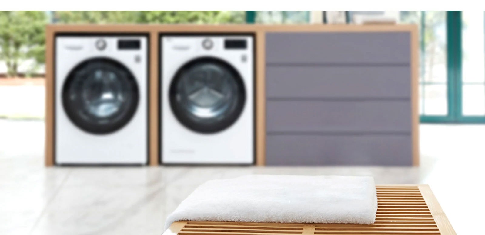 Background of the image is two washing machine front loading washers in a built in folding station slightly blurred with the LG ThinQ sitting on a towel on a table in the foreground.