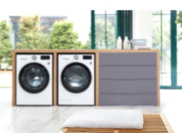 Background of the image is two washing machine front loading washers in a built in folding station slightly blurred with the LG ThinQ sitting on a towel on a table in the foreground.