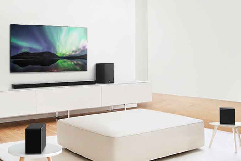 TV and sound bar in white living room with white sofa in the center. Speakers sit on both ends of the sofa.