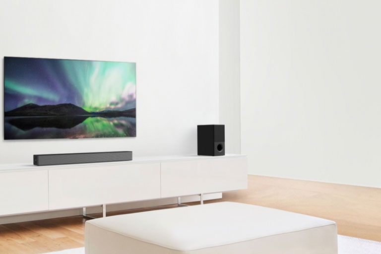 TV and sound bar in white living room with white sofa in the center. Speakers sit on both ends of the sofa.