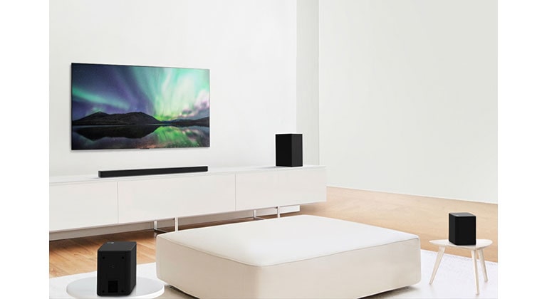 TV and sound bar in white living room with white sofa in the center. Speakers sit on both ends of the sofa.
