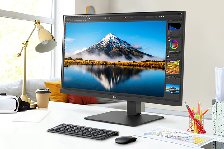 På skrivebordet er det en ultrafin skjerm som viser Photoshop-arbeid. Ved siden av skjermen er det en skrivebordslampe, en mus, papir og fargeblyanter.
