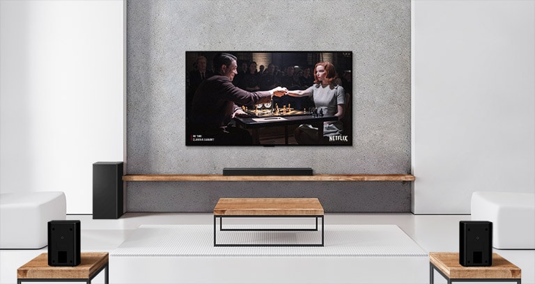 Et sett med to bakhøyttalere, subwoofer og en lydplanke og TV i en hvit stue. En kvinne og en mann spiller sjakk på en TV-skjerm. 