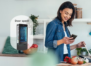 Et bilde av en kvinne som lager mat mens hun ser på telefonen og et kjøleskap med åpen dør.