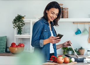 Et bilde av en kvinne som lager mat mens hun ser på telefonen og et kjøleskap med åpen dør.