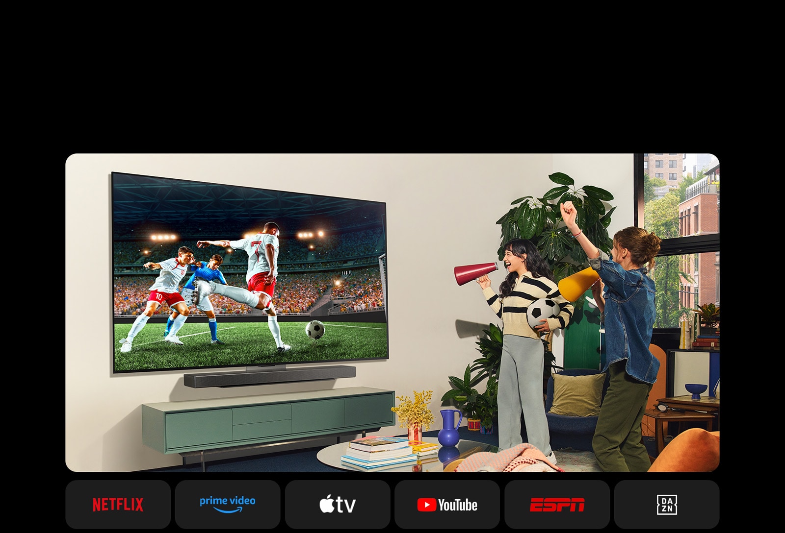 To kvinner ser på en fotballkamp i et koselig stueområde. En kvinne holder en fotball, og begge jubler for laget sitt med gule og røde høyttalere. De følgende logoene vises nedenfor. Netflix, Amazon Prime Video, Apple TV, Youtube, ESPN og DAZN.