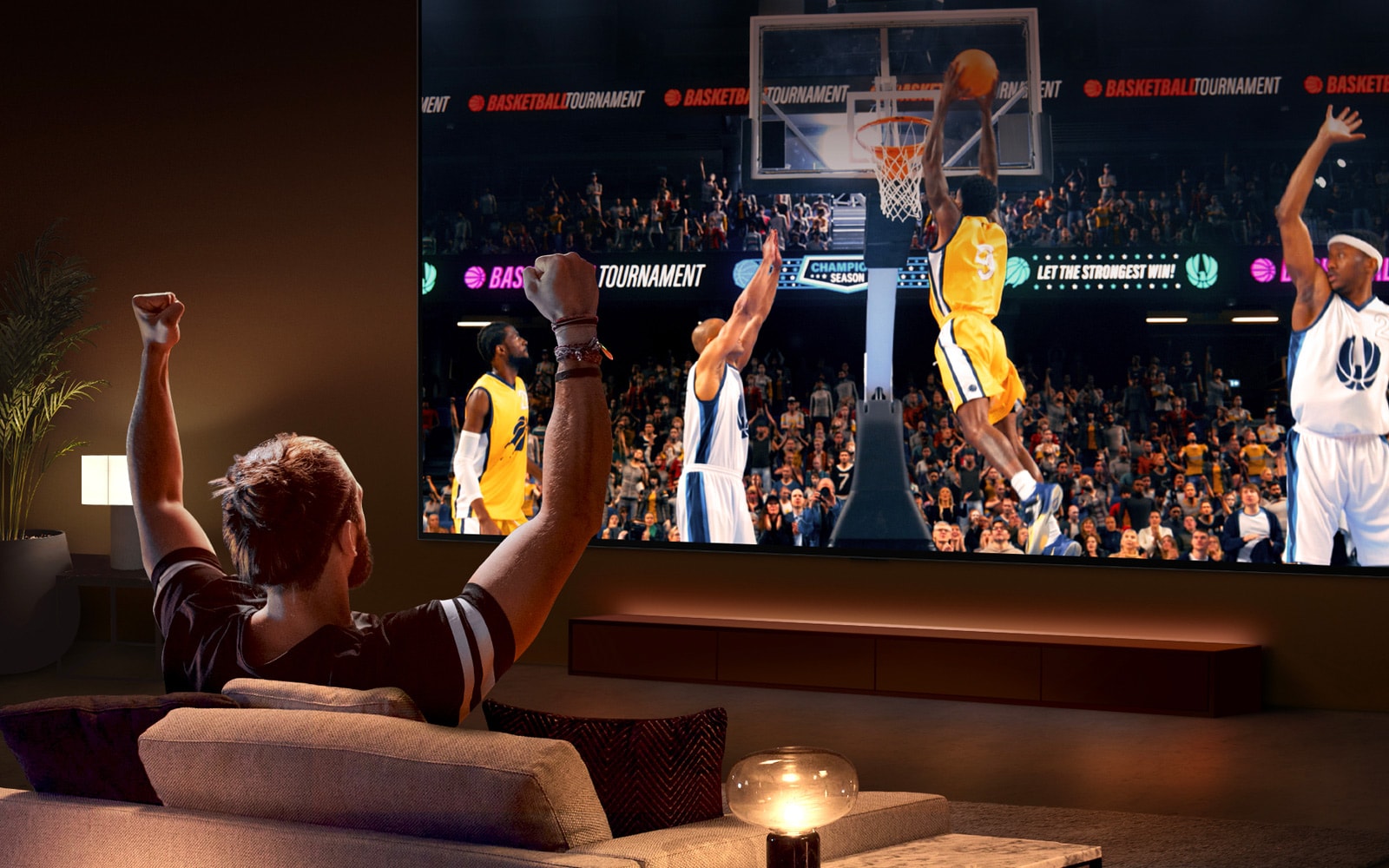 En mann sitter i en sofa foran en ultrastor LG-TV og viser en basketballkamp, med armene i været som om han jubler.