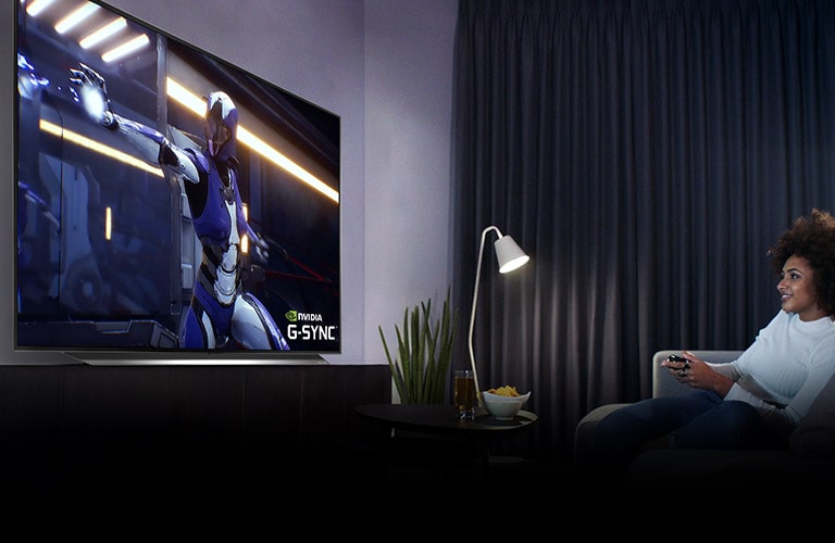 Kvinne som sitter i en sofa og holder på en joystick mens hun spiller et spill på en TV-skjerm