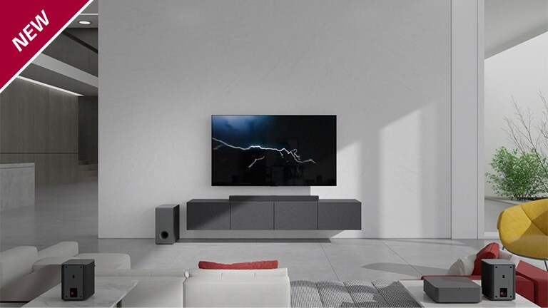 Lydplanken er plassert på et grått skap med en TV hengt på veggen i stuen. En trådløs subwoofer er plassert på gulvet på venstre side og sollyset kommer inn fra høyre side av bildet. En hvit- og rødfarget lang sofa står vendt mot TV-en og lydplanken. NYTT merke vises oppe i venstre hjørne.